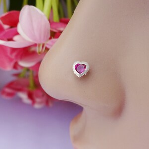 May include: A silver nose stud featuring a small, heart-shaped, vibrant pink gemstone set within a smooth, polished metal frame. The jewelry is positioned on the side of a nose, showcasing the heart-shaped design and the bright pink stone.