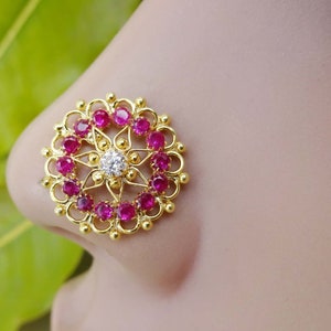 May include: Gold nose ring with a circular design featuring a central white stone surrounded by pink stones.