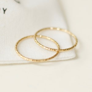 May include: Two gold-toned, textured, thin, metal rings on a white surface. The rings are stacked on top of each other. The background is a white fabric bag with the text "TESORE JEWELLERY" printed on it.