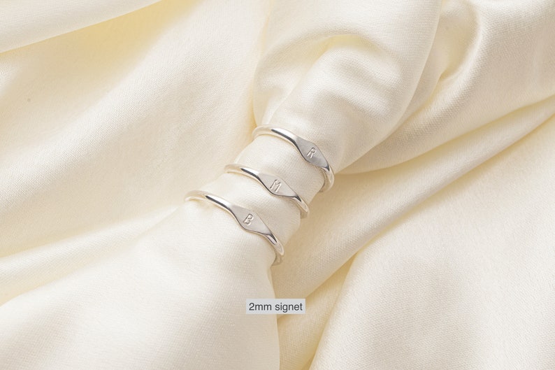 May include: Three silver signet rings with the letters B, M, and R engraved on them. The rings are stacked on top of each other and are sitting on a white fabric background. 2mm signet.