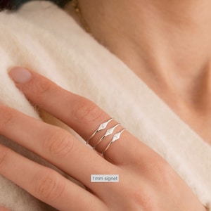 May include: Three silver signet rings stacked on a finger. The rings are thin and have a small, pointed diamond shape on the top.