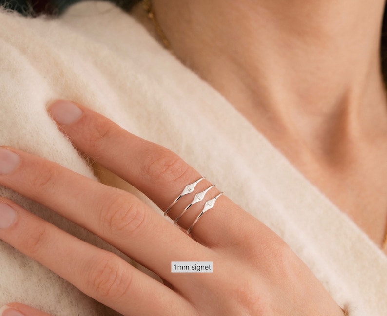 May include: Three silver signet rings stacked on a finger. The rings are thin and have a small, pointed diamond shape on the top.