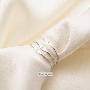 May include: Three silver signet rings with the letters "L", "R", and "F" engraved on them. The rings are stacked on top of each other and are on a white fabric background. 1 mm signet.