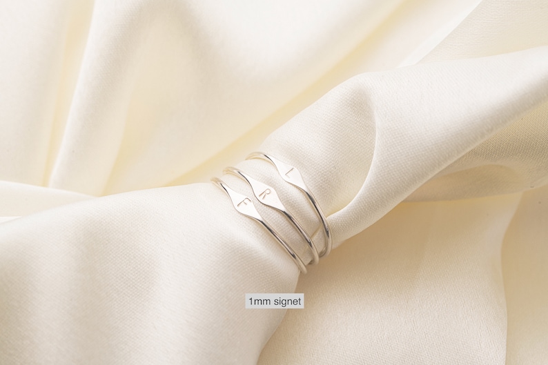 May include: Three silver signet rings with the letters "L", "R", and "F" engraved on them. The rings are stacked on top of each other and are on a white fabric background. 1 mm signet.