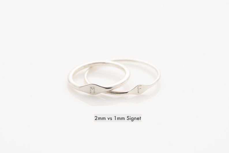 May include: Two silver signet rings with the letters "M" and "F" engraved on them. The rings are stacked on top of each other. The caption below the rings reads "2mm vs 1mm Signet".