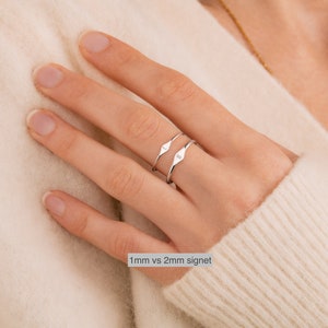 May include: Two silver signet rings on a finger. The top ring is 2mm wide and the bottom ring is 1mm wide. The text "1mm vs 2mm signet" is below the rings.