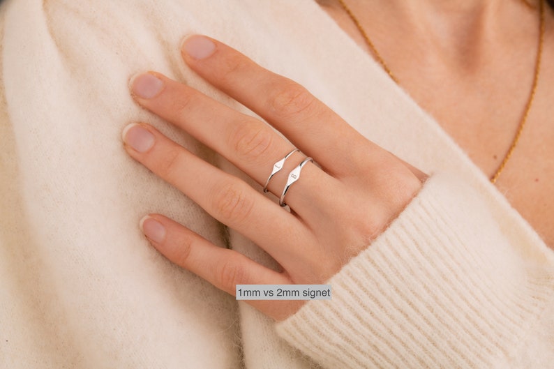May include: Two silver signet rings on a finger. The top ring is 2mm wide and the bottom ring is 1mm wide. The text "1mm vs 2mm signet" is below the rings.