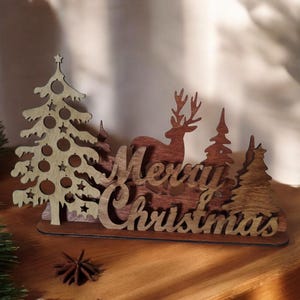 May include: Wooden Christmas decoration featuring a deer, trees, and the words "Merry Christmas." The design incorporates a light-colored Christmas tree with star and ball ornaments. The text and other elements are in a darker wood.