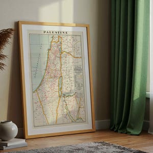 May include: A framed vintage map of Palestine, displayed indoors. The map, with the word "PALESTINE" at the top, features detailed geographical information in shades of beige, green, and red. The frame is light brown, and the map is leaning against a wall.