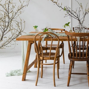 May include: A rustic wooden dining table set for a meal, surrounded by wooden chairs. The table is adorned with a sage green runner, glassware, and a vase of branches. The room has a bright, airy feel with white walls and floors.