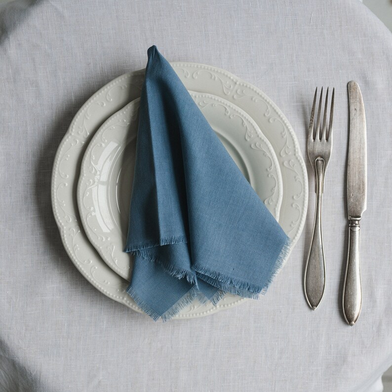 K&ouml;nnte beinhalten: Eine blaue Leinen-Serviette, die zu einem Dreieck gefaltet ist, liegt auf drei wei&szlig;en Tellern mit dekorativen R&auml;ndern. Die Teller befinden sich auf einer wei&szlig;en Tischdecke. Zwei silberne Besteckteile liegen rechts neben den Tellern.