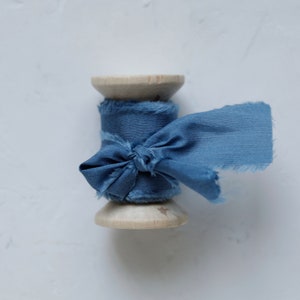 K&ouml;nnte beinhalten: Eine Spule mit blauem Band mit ausgefransten Kanten, zu einer Schleife gebunden. Das Band ist um eine Holzspule gewickelt.