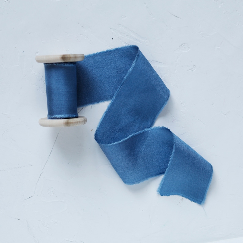 K&ouml;nnte beinhalten: Eine Spule mit blauem Seidenband mit einer Holzspule. Das Band ist abgewickelt und auf einer wei&szlig;en Oberfl&auml;che ausgelegt.