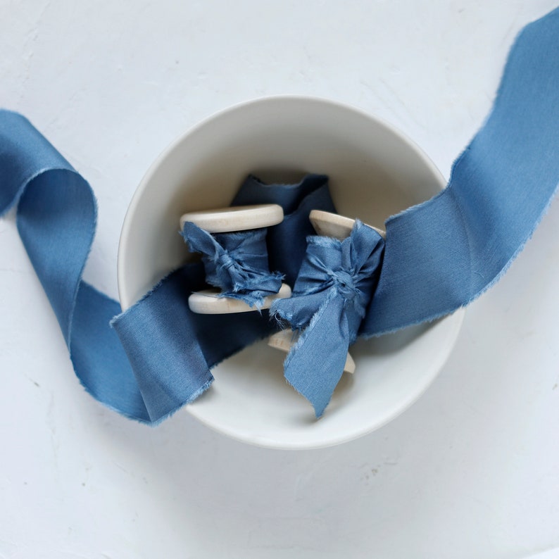 K&ouml;nnte beinhalten: Zwei Spulen mit blauem Seidenband in einer wei&szlig;en Sch&uuml;ssel. Das Band ist in einem weichen, staubigen Blau gehalten und hat einen leicht ausgefransten Rand.
