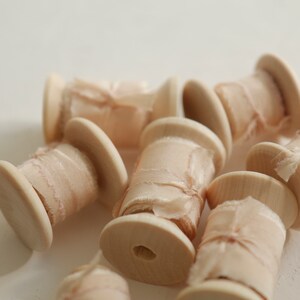 Pu&ograve; includere: Primo piano di diversi rocchetti di nastro di seta color blush avvolti attorno a rocchetti di legno. Il nastro &egrave; un tessuto delicato e trasparente con una superficie leggermente testurizzata.