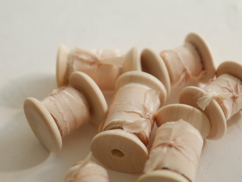 Pu&ograve; includere: Primo piano di diversi rocchetti di nastro di seta color blush avvolti attorno a rocchetti di legno. Il nastro &egrave; un tessuto delicato e trasparente con una superficie leggermente testurizzata.