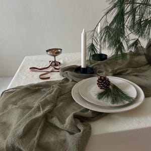 May include: A green linen table runner with a white tablecloth underneath. A white candle sits in a black holder, and a pine branch with a pine cone sits on a white plate.