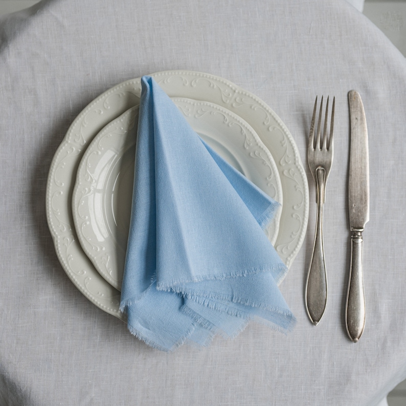 K&ouml;nnte beinhalten: Eine hellblaue Leinen-Serviette, die zu einem Dreieck gefaltet ist, liegt auf drei wei&szlig;en Tellern mit dekorativen R&auml;ndern. Zwei silberne Besteckteile liegen rechts neben den Tellern.