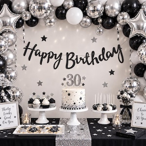 May include: A 30th birthday party display featuring a black and silver balloon arch with disco balls and star balloons. A cake with a 30 topper, cupcakes, and cookies are arranged on white stands. Two signs read Thirty Looks Good On You and Cheers to 30 Years.