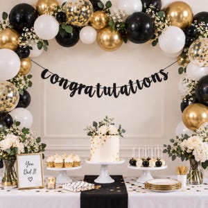 May include: A party decoration set featuring a black banner with the word congratulations, a white cake with gold accents, cupcakes, cake pops, and a balloon arch in black, white, and gold with clear balloons filled with gold confetti.