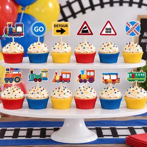 May include: A two-tiered white stand holding twelve cupcakes topped with train-themed paper decorations. The toppers include colorful locomotives, train cars, and road signs with text like GO, DETOUR, and RAIL CROSSING ROAD in red, blue, and yellow.