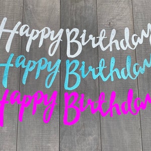 May include: Three glittery Happy Birthday banners in silver, light blue, and bright pink. Each banner features cursive script lettering with small holes at the top of each word for hanging. The letters have a textured, sparkling finish.