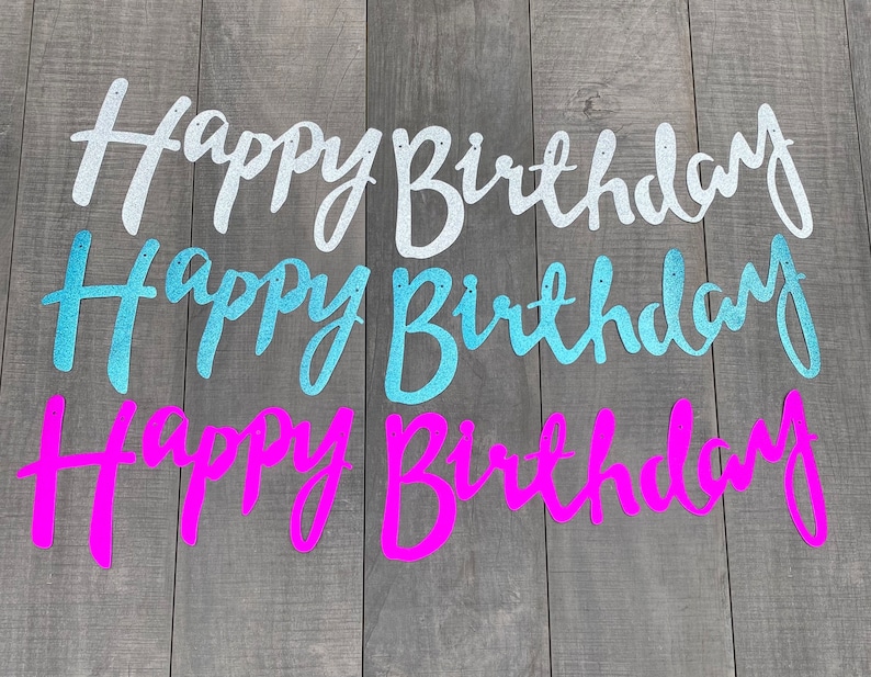 May include: Three glittery Happy Birthday banners in silver, light blue, and bright pink. Each banner features cursive script lettering with small holes at the top of each word for hanging. The letters have a textured, sparkling finish.