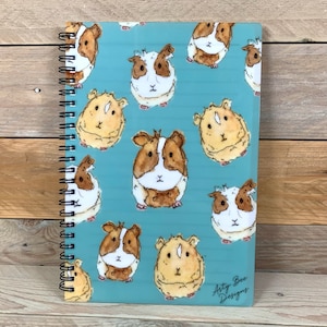 May include: A teal notebook with a spiral binding, decorated with watercolour illustrations of guinea pigs. The guinea pigs are in shades of brown, white, and tan. The notebook has a black spiral binding and the text "Arty Bee Designs" is visible.