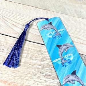 May include: A blue and white bookmark with a tassel. The bookmark features a repeating pattern of three dolphins jumping out of the water.