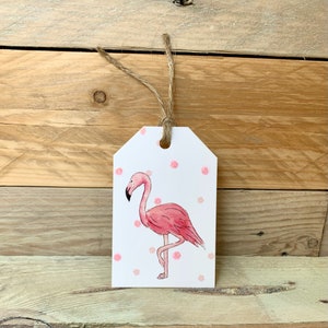 May include: A white gift tag featuring a pink flamingo illustration and pink polka dots. The tag is strung with natural twine. The background is a wooden surface.