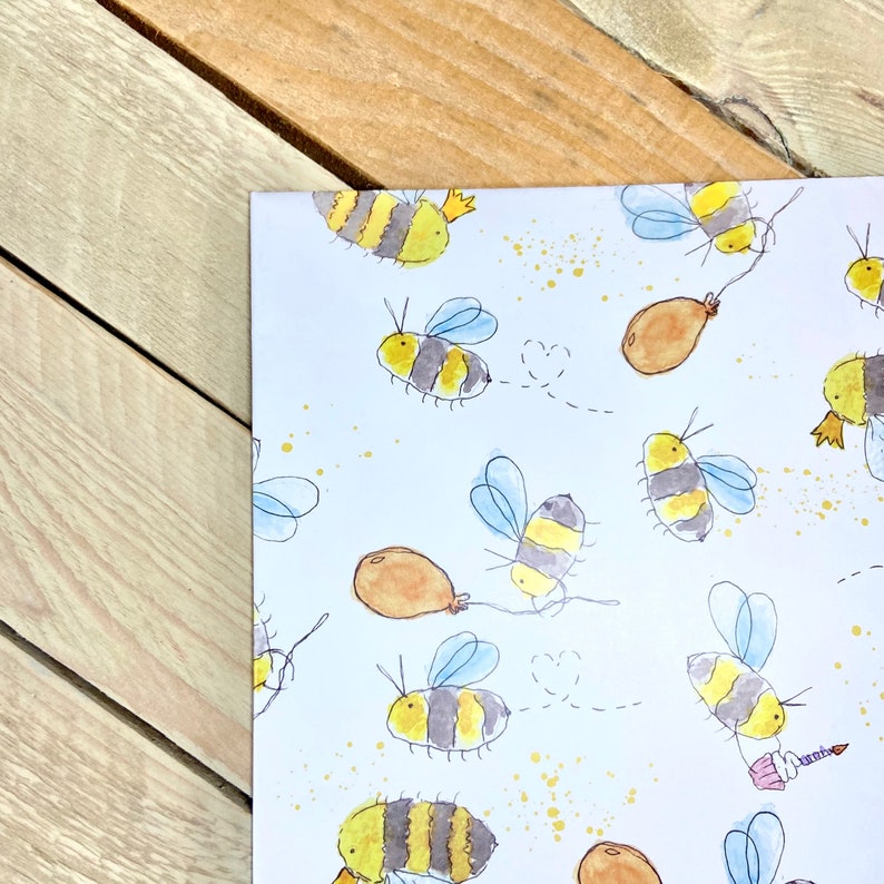 May include: White wrapping paper with a repeating pattern of yellow and black bees with brown balloons and gold glitter.