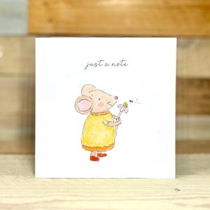 May include: A white greeting card with a watercolor illustration of a mouse holding a daisy. The mouse wears a yellow dress and red shoes. The text "just a note" is written in cursive at the top.