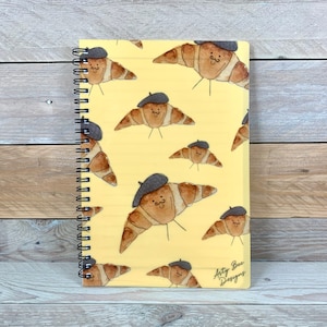 Könnte beinhalten: Ein gelber Spiralblock mit einem Muster aus braunen Croissants, die Baretts tragen und lächeln. Der Text "Arty Bee Designs" ist in der unteren rechten Ecke des Blocks gedruckt.