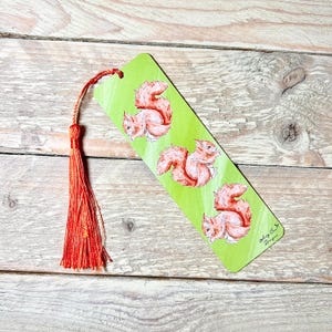 May include: A green bookmark with a red tassel features three watercolor illustrations of red squirrels. The bookmark has the text "Holly Red Designs" in the bottom right corner.