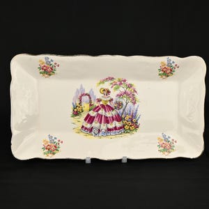 Könnte beinhalten: Ein rechteckiges Serviertablett in Weiß mit goldfarbenem Rand und floralem Design. In der Mitte des Tabletts steht eine Frau in einem rosa-weißen Kleid in einem Garten mit Blumen.