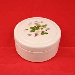 May include: White ceramic round box with a lid featuring a floral design of a strawberry plant with white flowers and red berries. The text "Fragaria vesca" is printed on the lid.