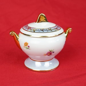 May include: A white porcelain sugar bowl with a lid, featuring gold accents and floral designs. The bowl has a rounded body, a pedestal base, and two gold handles. The lid is adorned with gold trim and a gold finial.