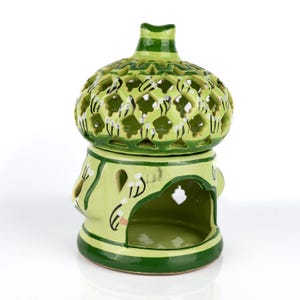 May include: A green ceramic oil warmer. The top section has a lattice design with cut-out patterns. The base features decorative cut-outs and painted floral accents. The warmer has a rounded shape and a small chimney.
