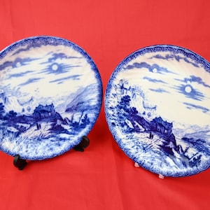 May include: Two blue and white decorative plates with a landscape design. The plates feature a detailed illustration of a building on a hill, mountains, and a moon in the sky. The plates have a scalloped edge.