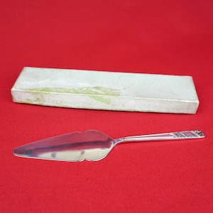 May include: A silver cake server with a decorative handle and a plain blade. The server is lying on a red surface next to a white box.
