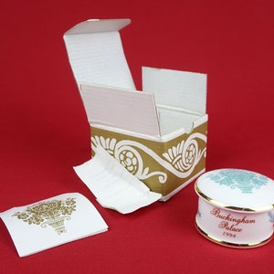 May include: A white porcelain trinket box with a gold trim and a blue and white floral design. The lid of the box has the text "Buckingham Palace 1994" printed on it. The box is sitting on a red surface and is next to a white box with a gold and white floral design.