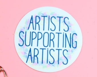 Op de afbeelding: Een ronde witte sticker met een blauw bloemenontwerp en de tekst "ARTISTS SUPPORTING ARTISTS" in blauwe letters.