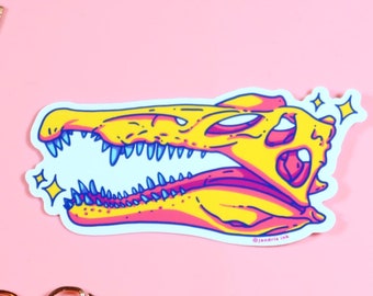 Op de afbeelding: Een gele en roze dinosaurus schedel sticker met een witte achtergrond en glinsterende details. De sticker is op een roze oppervlak.
