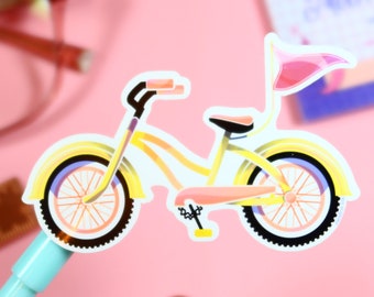 Op de afbeelding: Een gele fiets sticker met een roze vlag, een zwart zadel en zwarte wielen met witte spaken. De sticker zit op een blauwe pen.