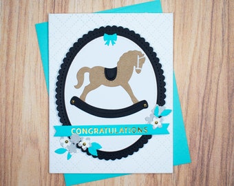Op de afbeelding: Een witte kaart met een zwart-wit silhouet van een schommelpaard in een zwarte cirkel met een gegolfde rand. De kaart heeft een turquoise blauw lint met het woord "CONGRATULATIONS" in gouden letters. De kaart is versierd met kleine witte bloemen en groene bladeren.