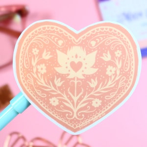 May include: A peach-colored heart-shaped sticker with a white floral design. The design features a stylized flower with a heart in the center. The sticker has a white border.
