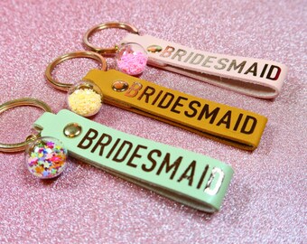 Op de afbeelding: Drie sleutelhangers met gouden sleutelringen en leren bandjes. Elk bandje heeft het woord "BRIDESMAID" in gouden letters gedrukt. De sleutelhangers hebben heldere glazen bedels gevuld met kleurrijk confetti.