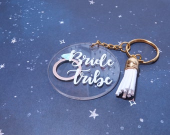Op de afbeelding: Een transparante acryl sleutelhanger met een witte kwast. De sleutelhanger heeft een ring met een diamant en de tekst "Bride Tribe".