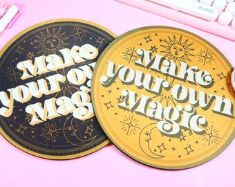 Op de afbeelding: Twee ronde muismatten met de tekst "Make your own Magic". Eén pad is zwart met gouden accenten, de andere is geel met gouden accenten. Beide hebben hemelse ontwerpen, waaronder zonnen, manen en sterren. Een roze toetsenbord en muis staan op de achtergrond.