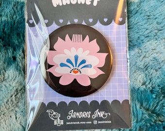 Op de afbeelding: Een ronde magneet met een bloemenontwerp in roze, blauw en wit op een zwarte achtergrond. De magneet is verpakt in een zwarte kaart met het woord "MAGNET" en het logo "Jandris Ink".